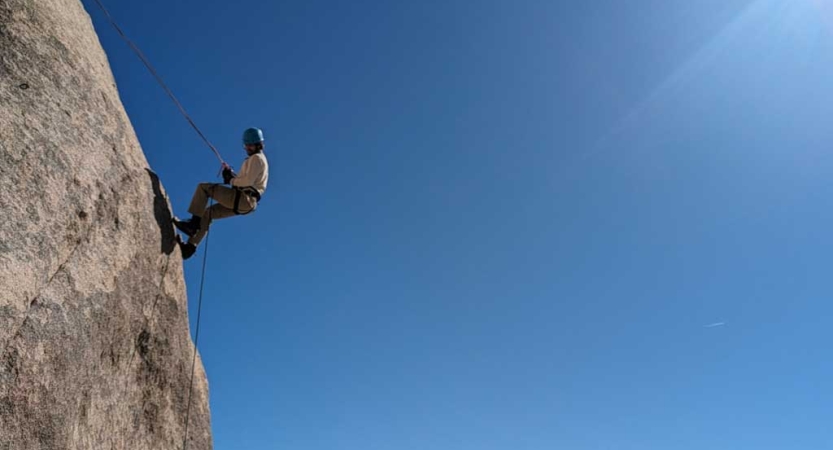 A person wearing safety gear is secured by ropes as they rappel from a rock wall. The sky is blue and cloudless behind them. 